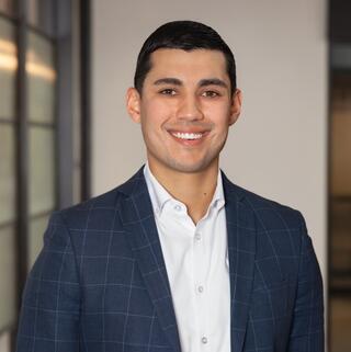 A young man smiles confidently in a smart navy blazer over a white shirt, standing in a well-lit, modern environment.