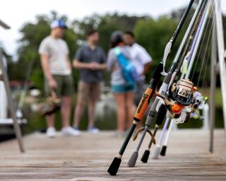 Fishing rods rest on a dock, with three people chatting in the background, surrounded by nature and a tranquil water scene.