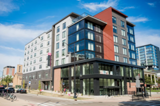 A modern multi-story building featuring a mix of glass and brick, with a bright blue sky above and a bustling street scene below.
