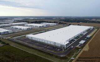 Aerial view of a large industrial complex with multiple warehouses, surrounded by open fields and distant structures on the horizon.