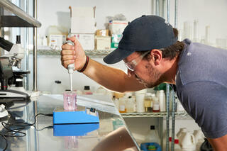 A researcher in a cap and goggles carefully uses a pipette to mix a pink solution on a lab countertop filled with equipment.
