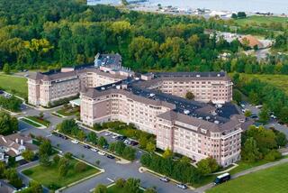 A large, multi-story building surrounded by lush trees and parking areas, with a glimpse of water in the background.