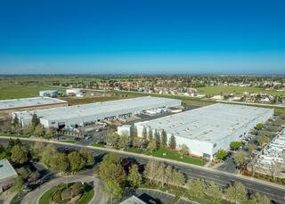 Large industrial complex with multiple warehouses and loading docks, surrounded by green fields and residential areas under a clear blue sky.