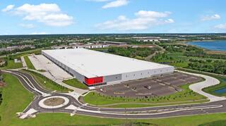A large, modern warehouse with a flat roof and red accents, surrounded by green landscaping and a spacious parking lot near a body of water.