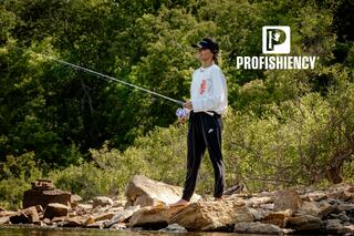A person stands on a rocky shoreline, fishing with a rod, dressed casually, surrounded by lush greenery and water.