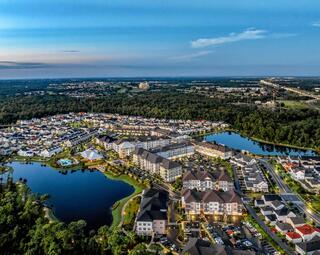 Aerial view of a suburban neighborhood with modern buildings, serene lakes, and greenery, featuring roads and amenities amidst a peaceful landscape.