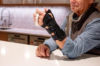 An older man with a wrist brace holds a cup, seated at a modern kitchen counter with a stylish design in the background.