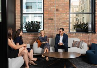 A group of four professionals engages in conversation in a stylish, modern office space with brick walls and large windows.