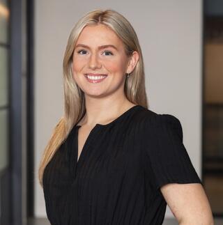 A smiling woman with long blonde hair stands confidently in a modern interior, wearing a black dress. The background features soft lighting.
