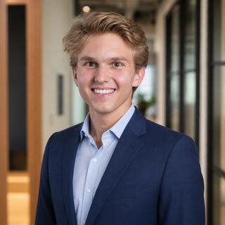A young man with tousled blond hair smiles confidently while wearing a blue suit and light shirt, set against a modern office backdrop.
