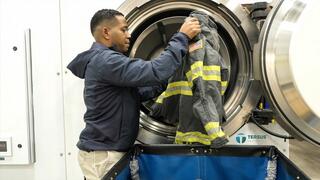 A person places a firefighter's jacket into a large washing machine in a clean, organized laundry facility.