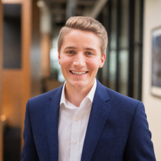 A young man in a blue blazer smiles warmly, set against a modern office backdrop with natural light filtering through.