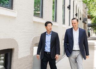 Two business professionals walk casually along a tree-lined city street, engaged in conversation, dressed in smart attire.