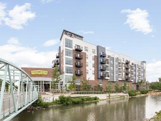 A modern multi-story building with balconies sits beside a river, featuring greenery and a pedestrian bridge in a bright, sunny atmosphere.