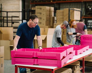 A group of individuals work in a warehouse, assembling or handling bright pink containers among stacks of cardboard boxes.