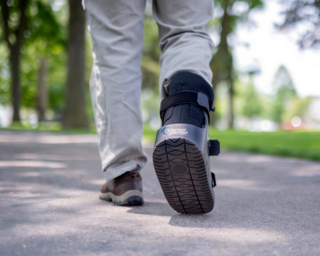 A person walks on a path, wearing a medical boot on one foot, with greenery and trees in the background.