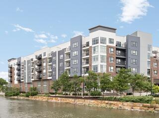 Modern multi-story apartment building beside a calm waterway, featuring greenery, balconies, and a clear blue sky.